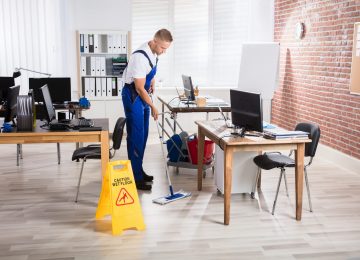Male Janitor Cleaning Floor With Caution Wet Floor Sign In Office